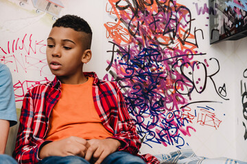 African american teen boy sitting in bed at home with drawings on wall
