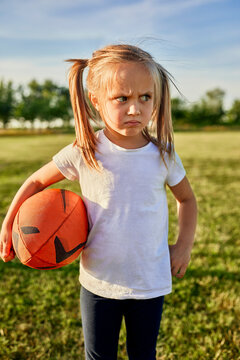 Displeased blond girl with rugby ball standing at sports field on sunny day