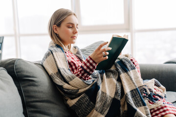 Young white woman wrapped in blanket reading book sitting on sofa