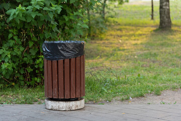 A wooden trash can stands in a green park on the sidewalk