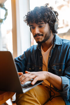 Young calm handsome indian curly man in headphones working laptop