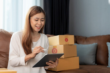 Asian female with small stock business owner holding digital tablet and retail package parcel boxes checking commercial shipping delivery order on smartphone using mobile app technology.