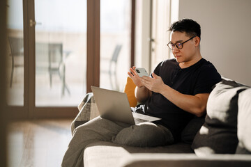 Adult brunette asian man using cellphone and laptop at home