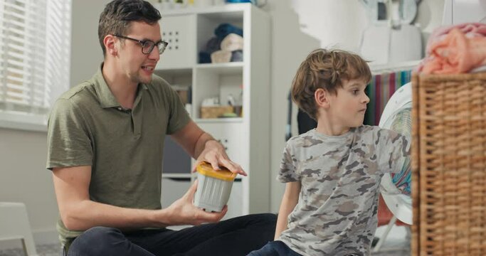 Father spends time with young children in laundry room, bathroom, performs household duties while on paternity leave, takes care of son, shows boy how to operate washing machine turn on program.