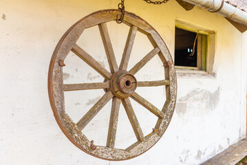 Old wooden carriage wheel with metal fittings hangs on the white wall. Outdoor natural day light.