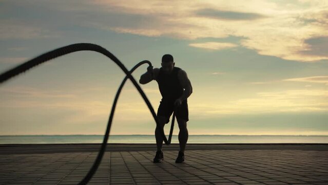 Bodybuilder training outdoors doing an exercise with fighting ropes against the backdrop of sunrise. Training lifestyle.Slow Motion