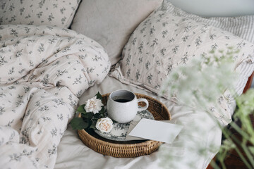 Breakfast in bed. Bedroom interior. Cup of coffee, rose flowers on porcelain plate. Wicker tray. Blank greeting card, invitation mockup. Llinen bedding. Vintage design. Blurred floral foreground.