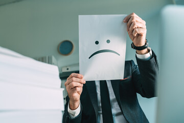 Unhappy businessman holding paper with frowning emoticon in office interior - obrazy, fototapety, plakaty