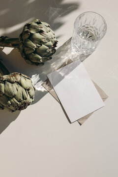 Summer stationery. Food still life scene. Glass of water, green artichoke vegetable in light, shadows. Beige sunny table background. Blank paper card, invitation mockup scene. Vertical flat lay, top.