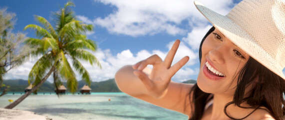 travel, tourism and summer vacation concept - happy smiling woman in straw hat showing peace gesture over tropical beach background in french polynesia