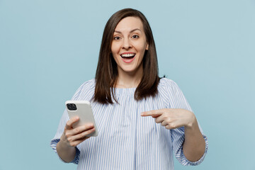 Young happy shocked fun woman she 20s in casual blouse hold in hand use point index finge on mobile cell phone isolated on pastel plain light blue background studio portrait. People lifestyle concept.