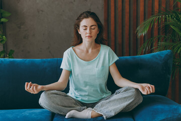 Young spiritual woman she 20s wear casual clothes mint t-shirt hold spreading hands in yoga om aum gesture relax meditate try to calm down sit on blue sofa indoor rest at home in own room apartment