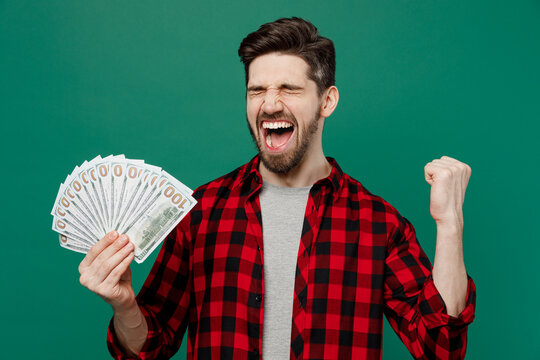 Young Happy Smiling Man He 20s Wearing Red Shirt Grey T-shirt Holding Fan Of Cash Money In Dollar Banknotes Do Winner Gesture Isolated On Plain Dark Green Background Studio. People Lifestyle Concept.