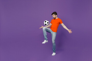 Fototapeta na wymiar Full body young cheerful excited fun fan man he 20s wear orange t-shirt cheer up support football sport team juggling soccer ball on knee watch tv live stream isolated on plain dark purple background.