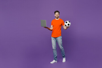 Full body young fan man he wear orange t-shirt cheer up support football sport team hold soccer ball watch tv live stream use betting on laptop pc computer isolated on plain dark purple background