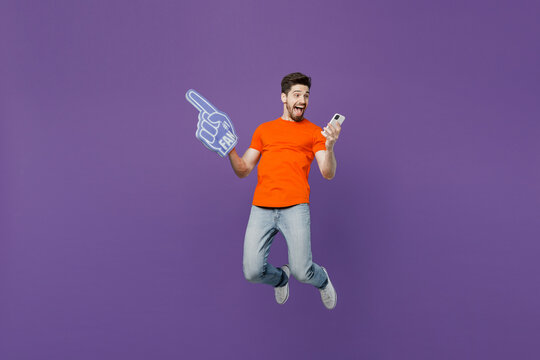 Full body young fan man he wear orange t-shirt cheer up support football sport team hold mobile cell phone fan foam glove finger up watch tv live stream jump isolated on plain dark purple background.