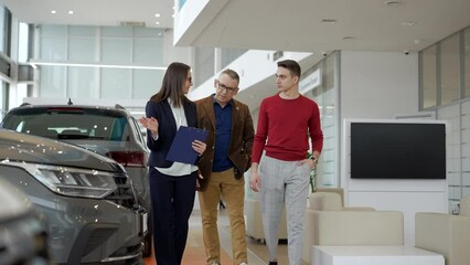 Two family people are talking to female agent and walking in car dealership office spbd. 4k Elderly father, young son walk in sales room and talk with woman consultant, study car model and make - Powered by Adobe