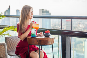 Beautiful young woman with cocktails rests at luxury rooftop restaurant. Elegant female lady in red dress with mocktails at sky bar terrace looking at modern city skyline. Skyscrapers on background.