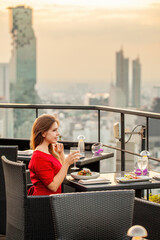 Beautiful young woman with cocktail rests at luxury rooftop restaurant at sunset. Elegant female in...