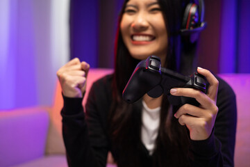 Playing video game. Young asian pretty woman with headphone sitting on sofa holding joystick in living room. Happy female Professional Streamer chinese playing game online in dark room neon light.