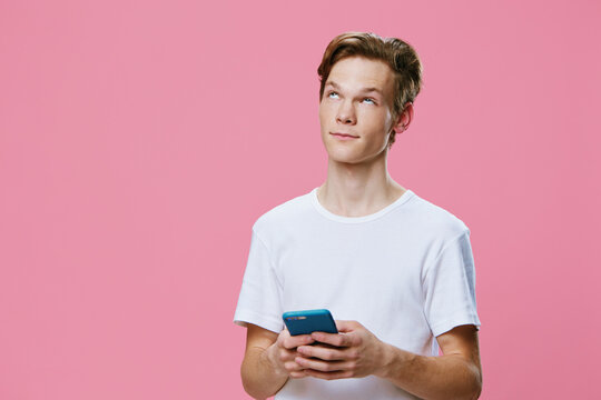 a handsome guy in a white t-shirt looks up thoughtfully while standing with a phone in his hands on a pink background