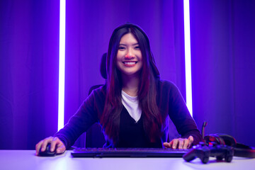 Fototapeta na wymiar Playing video game. Young asian pretty woman sitting on chair with computer pc in living room. Happy female Professional Streamer chinese wearing hoodie playing game online in dark room neon light.