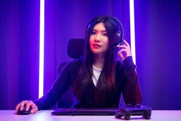 Professional gamer. Young asian pretty woman sitting on chair with computer pc in living room. Happy female Professional Streamer chinese wearing headphone playing game online in dark room neon light