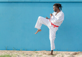 a young african american man training taekwondo outdoors kicking on a blue background