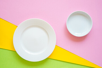 Ein leerer weißer Teller und eine Schüssel auf einem rosa, gelb und grünen Hintergrund....