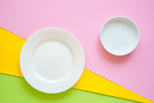 Ein leerer wei&szlig;er Teller und eine Sch&uuml;ssel auf einem rosa, gelb und gr&uuml;nen Hintergrund. Draufsicht, modern.