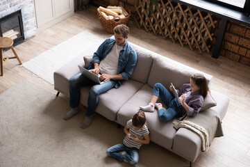 Young family of three holding and using different electronic devices while sitting on sofa in the living room - obrazy, fototapety, plakaty