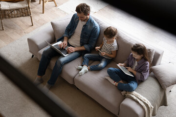 Young family of three holding and using different electronic devices while sitting on sofa in the...