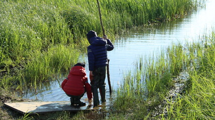 A boy in a red jacket and a blue hat stands on the swampy bank of the river