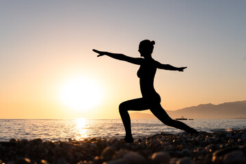 Fototapeta na wymiar Yoga wellness retreat class on morning sunrise beach landscape. Silhouette of girl standing in warrior pose meditation vertical background.