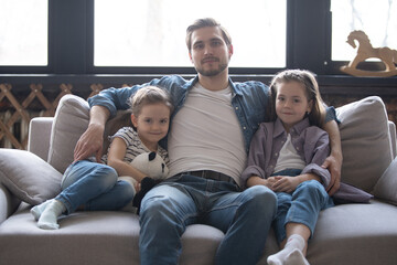 Little daughters sitting next to daddy at home smile look at camera. Celebrating Happy Father Day concept