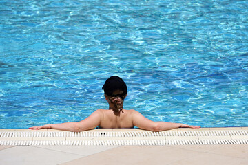 Young woman in baseball cap tanning in transparent water leaning on edge of swimming pool, rear view. Summer leisure and beach vacation