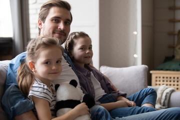 Fototapeta premium Little daughters sitting next to daddy at home smile look at camera. Celebrating Happy Father Day concept