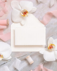 Handmade paper card and envelope near white orchid flowers and silk ribbons on marble, mockup