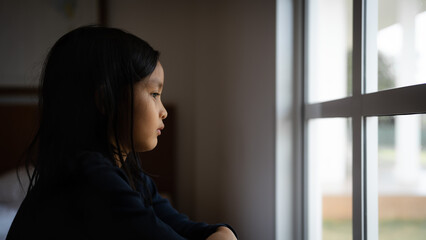 Fototapeta na wymiar Sad Asian little girl is looking outside window and feeling lonely in her bedroom, concept of depressed, unhappy, and help support need, autism, mental health.