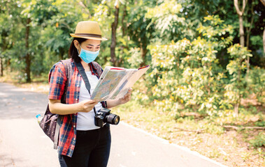 Asian woman tourism traveling in a beautiful green park wearing protective mask with camera and backpack and hold map ,Asian women looking map during travel in forest ,Outdoor new normal social distan