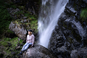 Mature female woman sitting on a wet rock in front of a wild waterfall in northern Sweden.