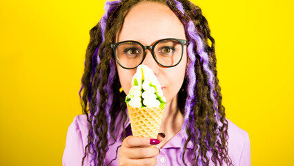 Young beautiful woman in glasses with at ice cream, yellow background. Portrait of happy female with dreadlocks in anticipation enjoying sundae.