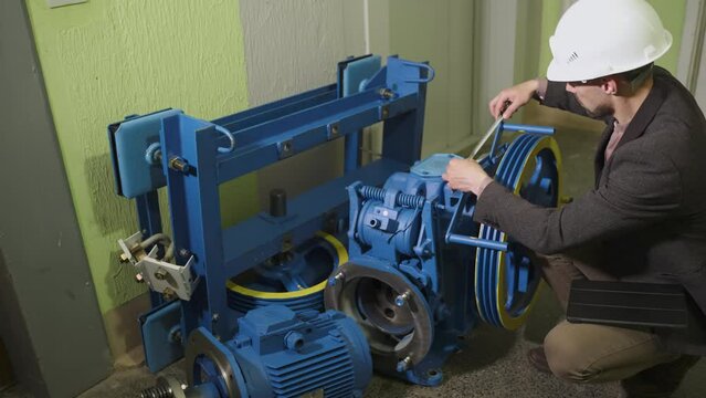 Engineer in helmet measures parameters of new blue, yellow elevator electric motor with measuring tape before installation, enters data into tablet. Controller checks the elevator equipment.