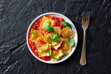 Ravioli with tomato sauce and fresh basil leaves on a plate, overhead flat lay shot on a black slate background, with a fork