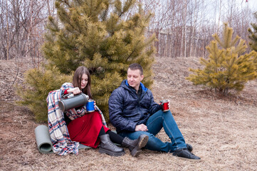 Naklejka na ściany i meble A couple in a spring park: a man and a girl sit near a fir tree and drink hot tea from a thermos