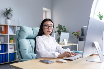 Portrait of successful office worker Asian woman, looking at camera smiling, business woman in glasses working and typing on computer.