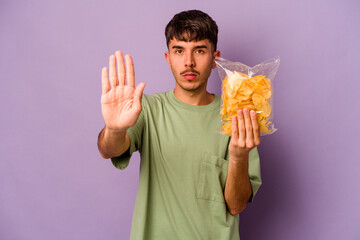Young hispanic man holding crisps isolated on purple background standing with outstretched hand showing stop sign, preventing you. - obrazy, fototapety, plakaty