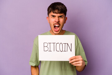 Young hispanic man holding bitcoin placard isolated on purple background screaming very angry and aggressive.