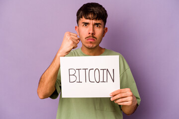 Young hispanic man holding bitcoin placard isolated on purple background showing fist to camera, aggressive facial expression.