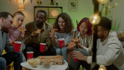 Diverse group of students eating pizza drinking and laughing enjoying indoor party in apartment at night. Youth lifestyle and communication concept.